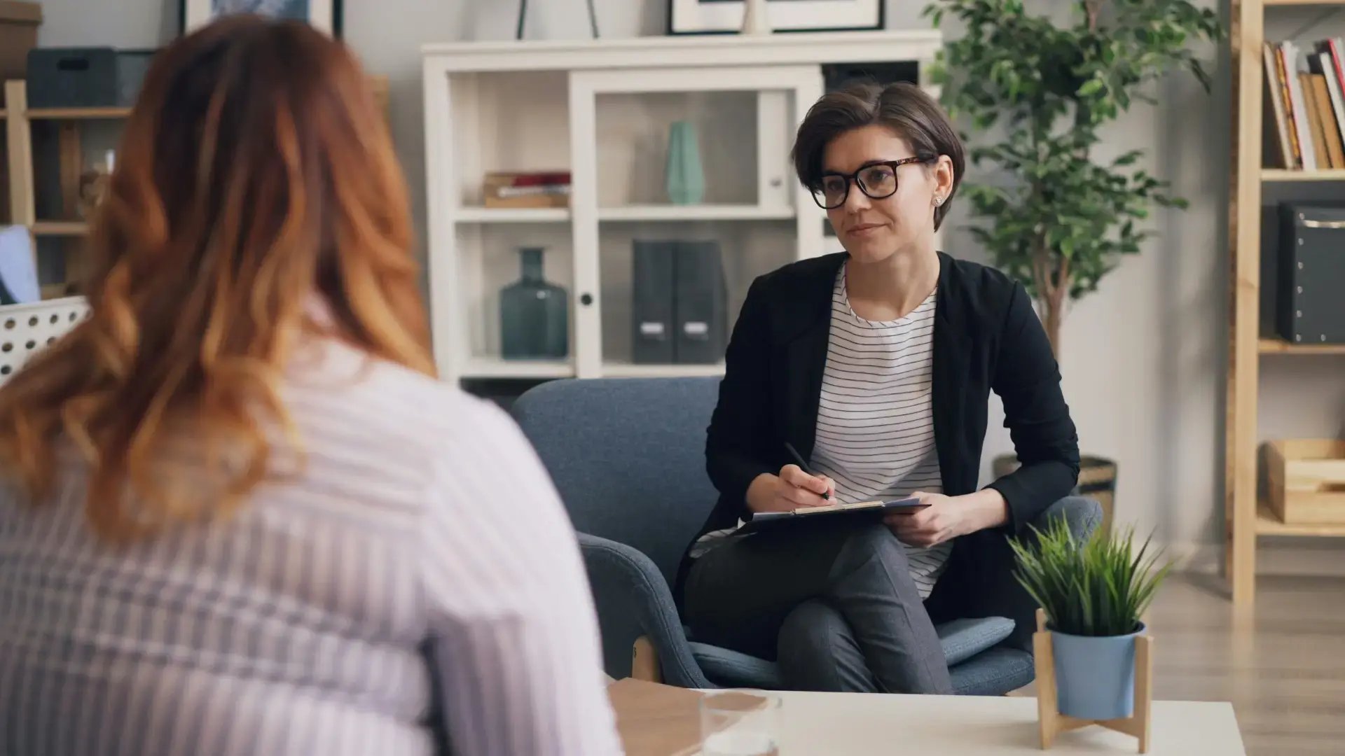 A therapist with short brown hair and glasses sits across from a client in a therapy office, holding a clipboard and listening attentively during a session. Trauma Therapist Institute