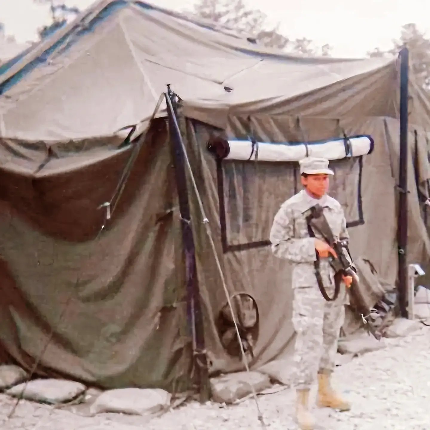 A soldier in Army combat uniform holding a rifle while standing outside a large canvas field tent at a military encampment