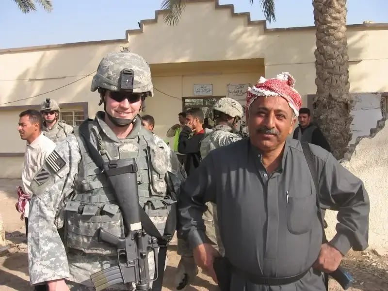A U.S. Army soldier in full combat gear smiling alongside an Iraqi man in a traditional keffiyeh outside a building during a deployment in Iraq