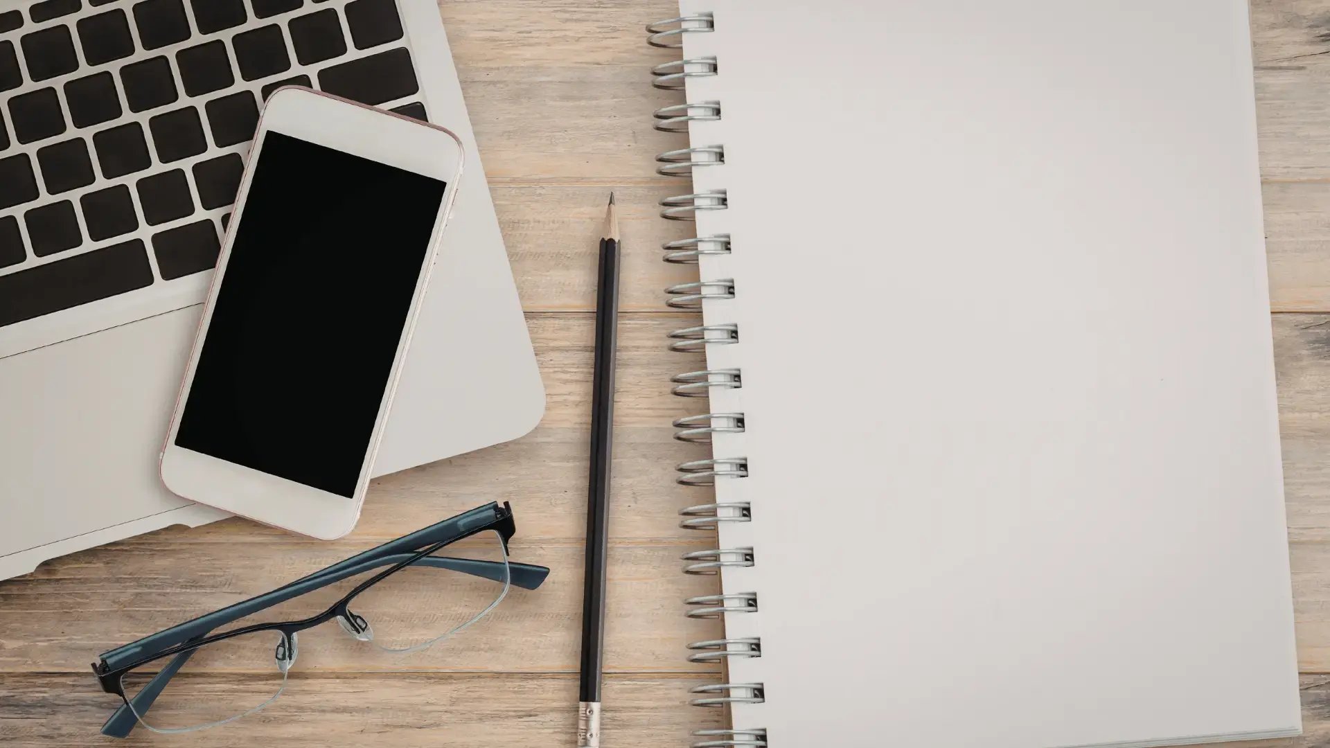 Overhead view of a workspace with a laptop, phone, glasses, pencil, and open notebook on a wooden desk