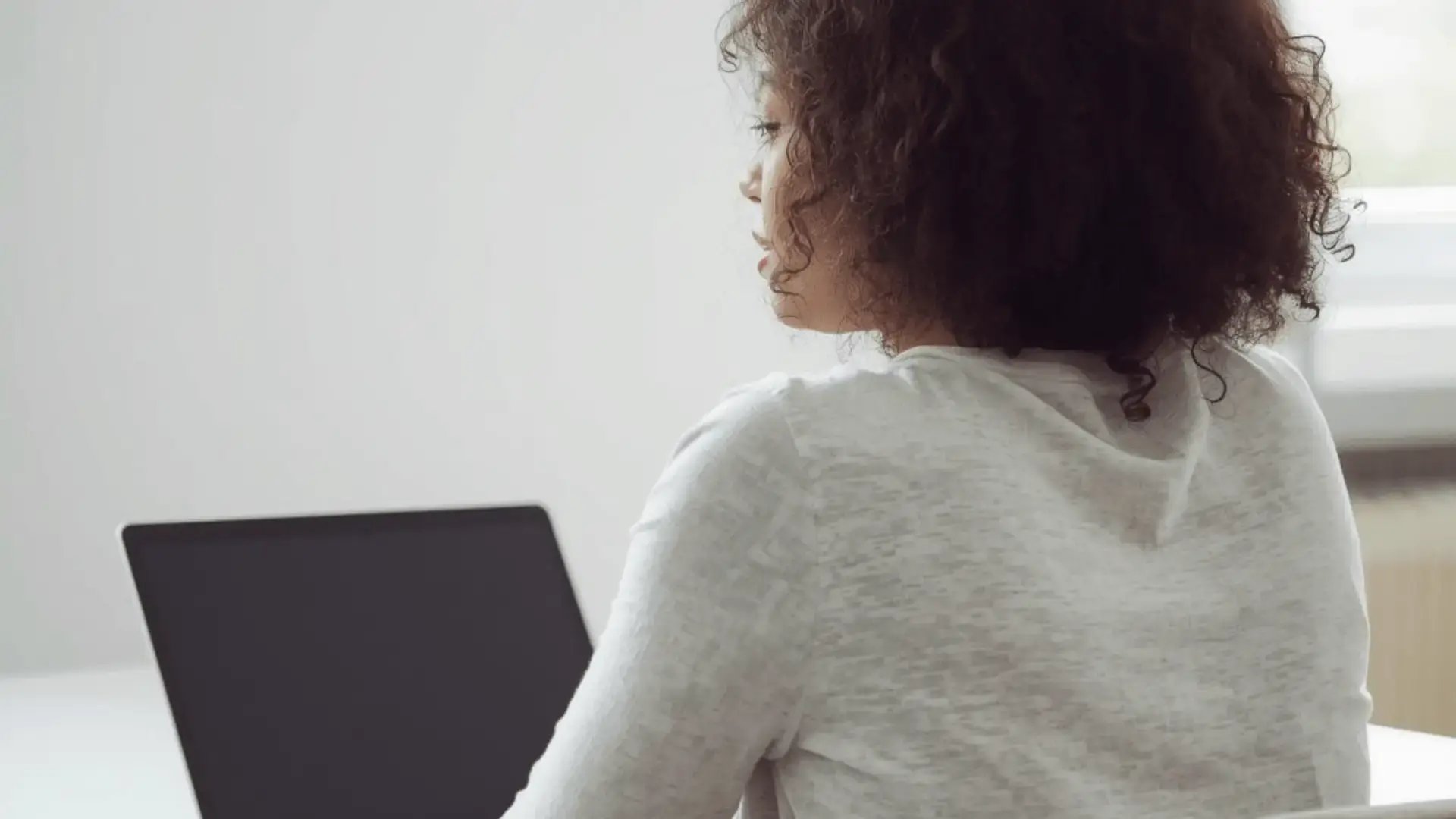 Woman seen from behind working at a laptop in a bright, minimally decorated workspace with natural light Trauma Therapist Institute
