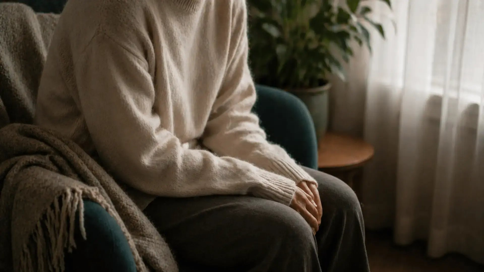 A person sits alone in a soft chair near a window with shoulders rounded forward and posture collapsed inward, body wrapped in an oversized knit sweater, illustrating the felt experience of dorsal vagal shutdown — the nervous system's freeze response of withdrawal and disconnection.
